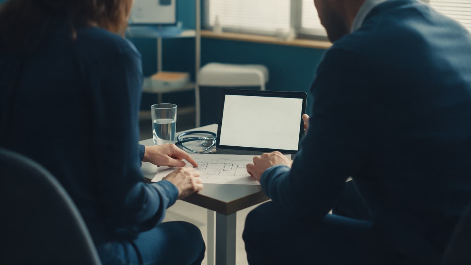 Two adults seen from behind at a consulting-room table, one pointing at a printed plan beside a tablet